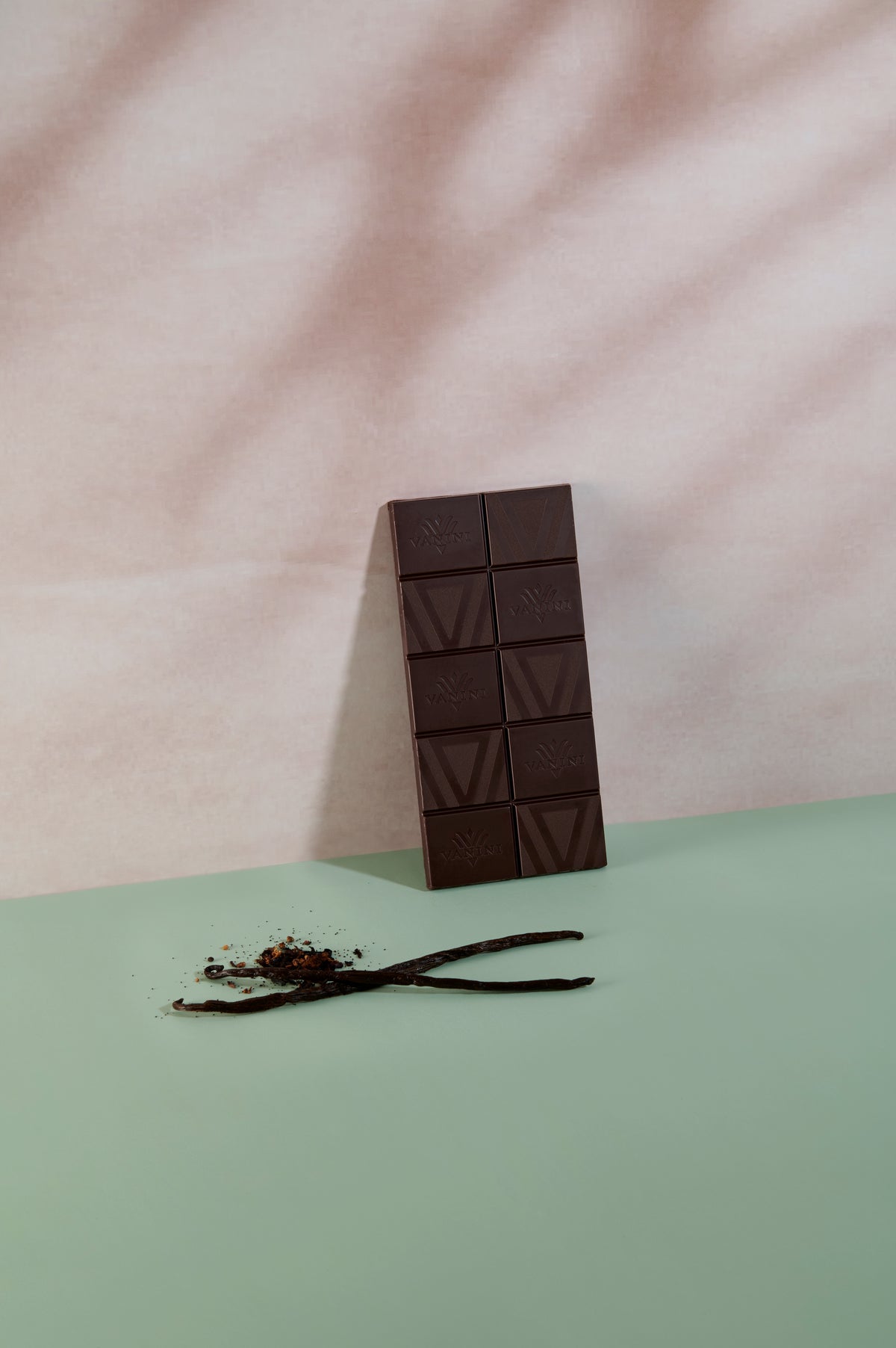 Tavoletta di cioccolato fondente Vanini Giamaica 80% con bacche di vaniglia macinate, fotografata accanto a baccelli di vaniglia e scaglie di cacao su sfondo dai toni verde salvia e rosa.