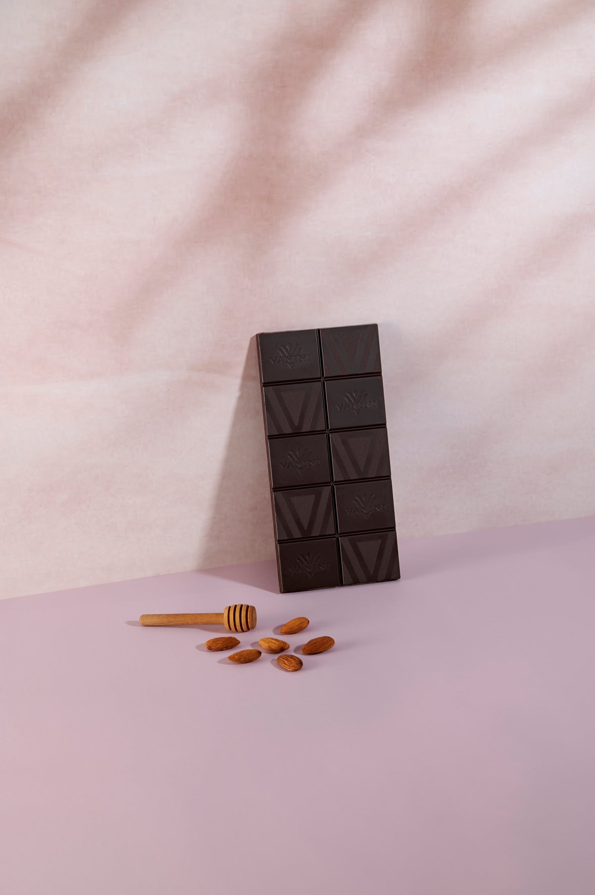 Tavoletta di cioccolato fondente Vanini Ecuador 80% senza confezione, disposta verticalmente su una superficie rosa tenue. In primo piano sono presenti mandorle intere e un spargimiele in legno, ingredienti che richiamano le note di miele e mandorla della ricetta. La finitura scura del cioccolato e l'incisione del logo Vanini risaltano sullo sfondo luminoso e minimale.