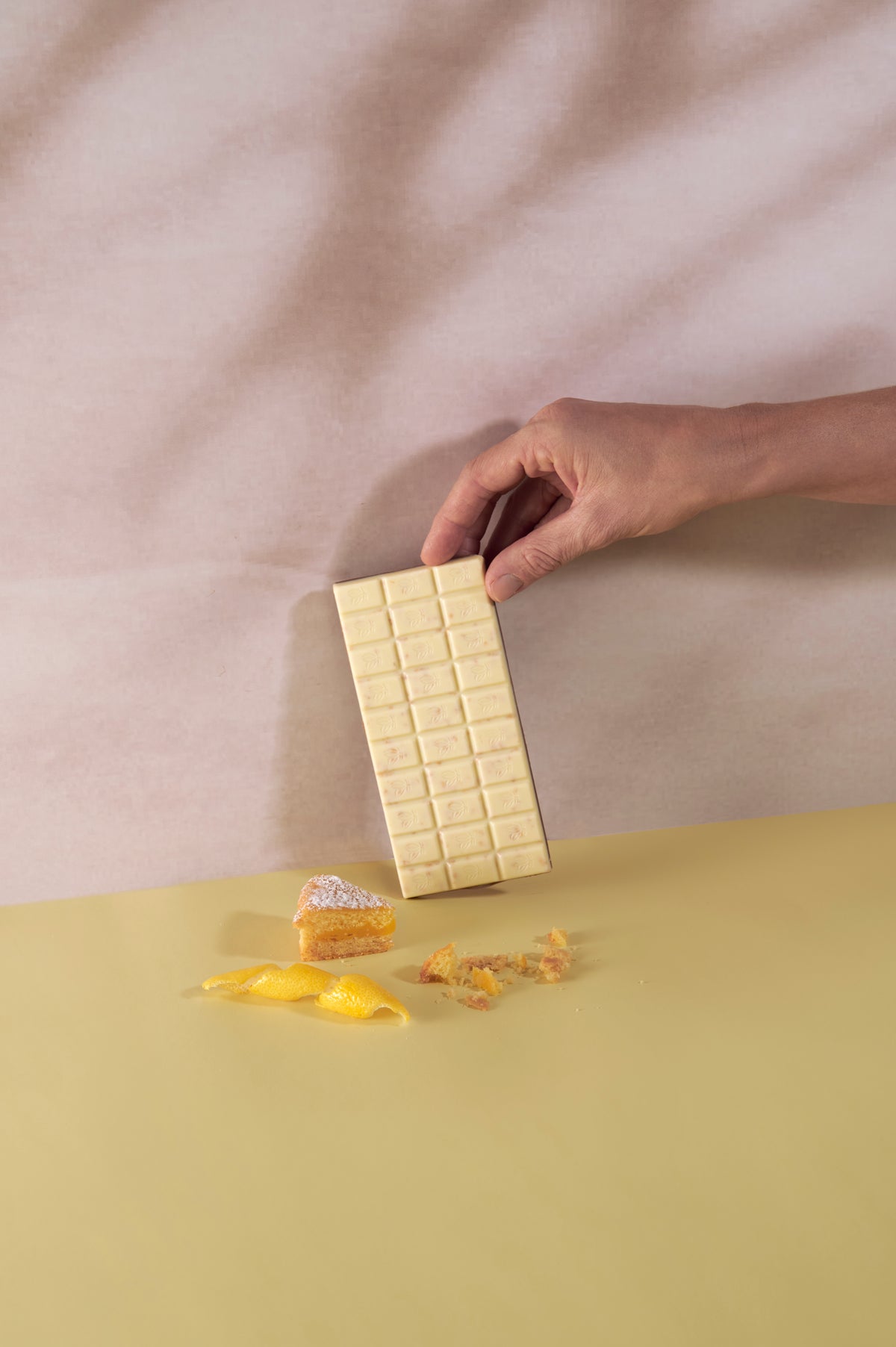 Una mano sorregge una tavoletta di cioccolato bianco Vanini Tasting Experience Double Torta al Limone. Sul piano sottostante sono presenti una fetta di torta al limone, scorza di limone e briciole di dolce su sfondo giallo tenue.