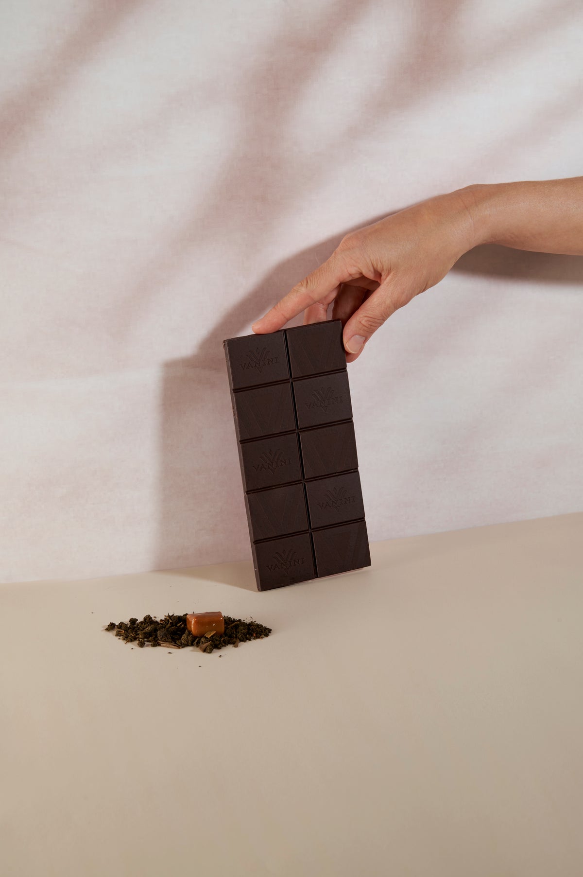 Mano che solleva una tavoletta di cioccolato fondente Vanini Madagascar 80% al tè chai con cristalli di caramello. La tavoletta, composta da dieci quadratini incisi con il logo Vanini, è fotografata in posizione verticale su una superficie beige dallo stile minimale. In primo piano sono disposte foglie di tè chai sfuse e un cristallo di caramello, ingredienti che richiamano il profilo aromatico speziato della ricetta. Lo sfondo chiaro con ombre morbide valorizza il colore intenso del cioccolato e l'eleganza