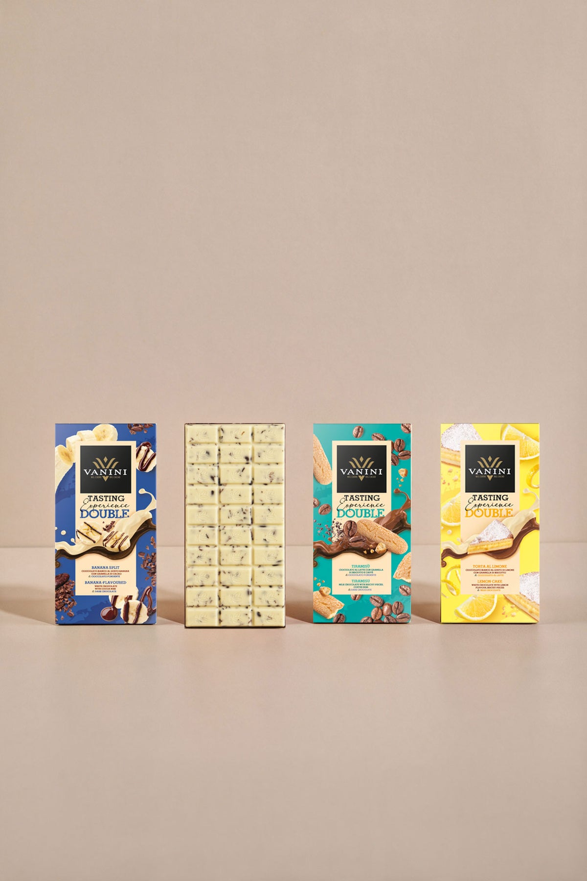 Assortimento Vanini Double Tasting Experience con quattro tavolette ispirate a celebri dessert internazionali. Al centro della composizione è esposta una tavoletta di cioccolato bianco aperta, decorata con inclusioni visibili, accanto alle confezioni Banana Split, Tiramisù e Torta al Limone. Le grafiche colorate e gli ingredienti illustrati, come banane, biscotti, chicchi di caffè e limoni, valorizzano la varietà dei gusti e l’esperienza di degustazione proposta dalla collezione.