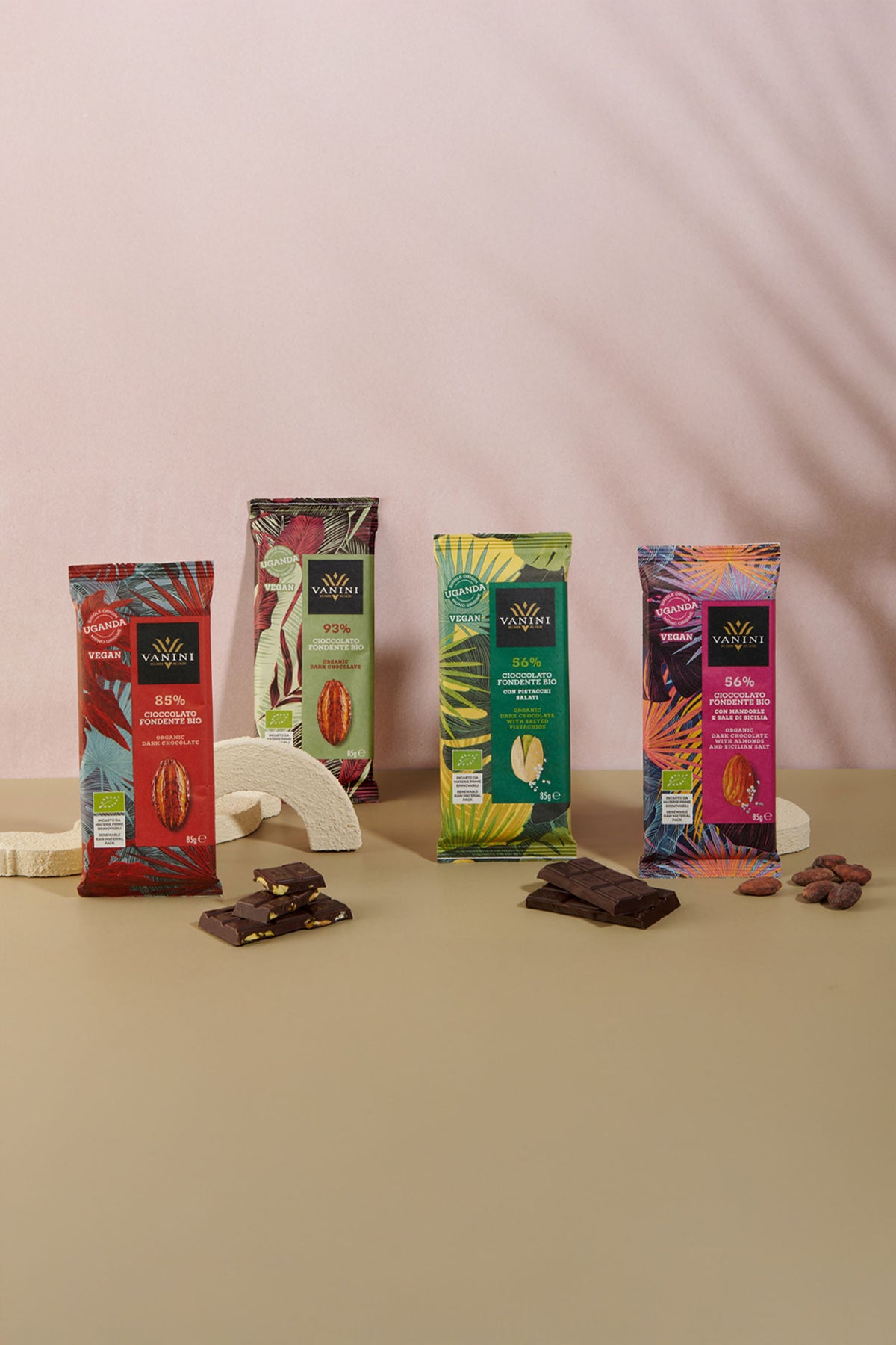 Selezione di quattro tavolette di cioccolato biologico Vanini Uganda disposte su sfondo beige e rosa cipria. La collezione comprende cioccolato fondente bio 85%, fondente bio 93%, fondente bio 56% con pistacchio e fondente bio 56% con mandorle e sale di Sicilia. In primo piano sono visibili pezzi di cioccolato, fave di cacao, pistacchi e elementi decorativi color avorio che valorizzano il packaging tropicale ispirato alle foreste dell'Uganda.