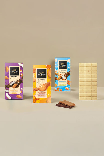 Selezione di tavolette Vanini Tasting Experience con esposizione di una tavoletta di cioccolato bianco aperta accanto alle confezioni della collezione. La composizione presenta gusti arricchiti da inclusioni selezionate come cocco, mandorle, granella di biscotto, popcorn caramellati, sale e pistacchio salato. Il design moderno e colorato delle confezioni è valorizzato da uno sfondo beige elegante che mette in risalto le diverse specialità di cioccolato.