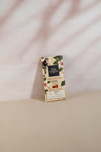 Tavoletta di cioccolato fondente Vanini Madagascar 80% con tè chai e cristalli di caramello, confezione illustrata con fiori tropicali e lemure del Madagascar. Il prodotto è fotografato in verticale su una superficie beige con sfondo chiaro dalle ombre morbide, in uno stile elegante e minimale.