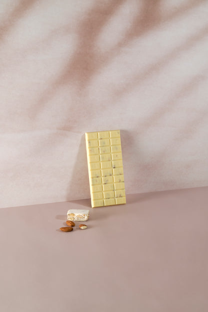 Tavoletta di cioccolato bianco Vanini Tasting Experience gusto torrone con inclusioni di mandorle, affiancata da un pezzo di torrone tradizionale e mandorle intere su sfondo rosa delicato.