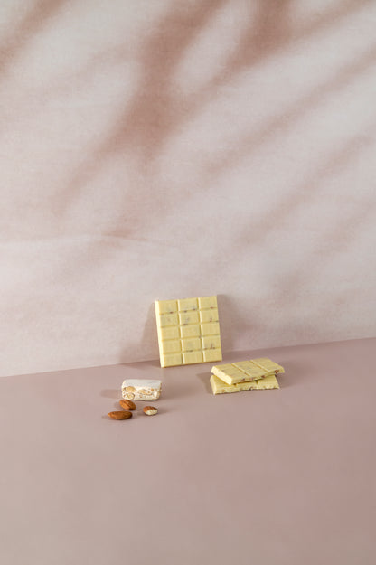 Tavoletta di cioccolato bianco Vanini Tasting Experience gusto torrone divisa a pezzi, con inclusioni di mandorle, affiancata da un pezzo di torrone tradizionale e mandorle intere su sfondo rosa delicato.