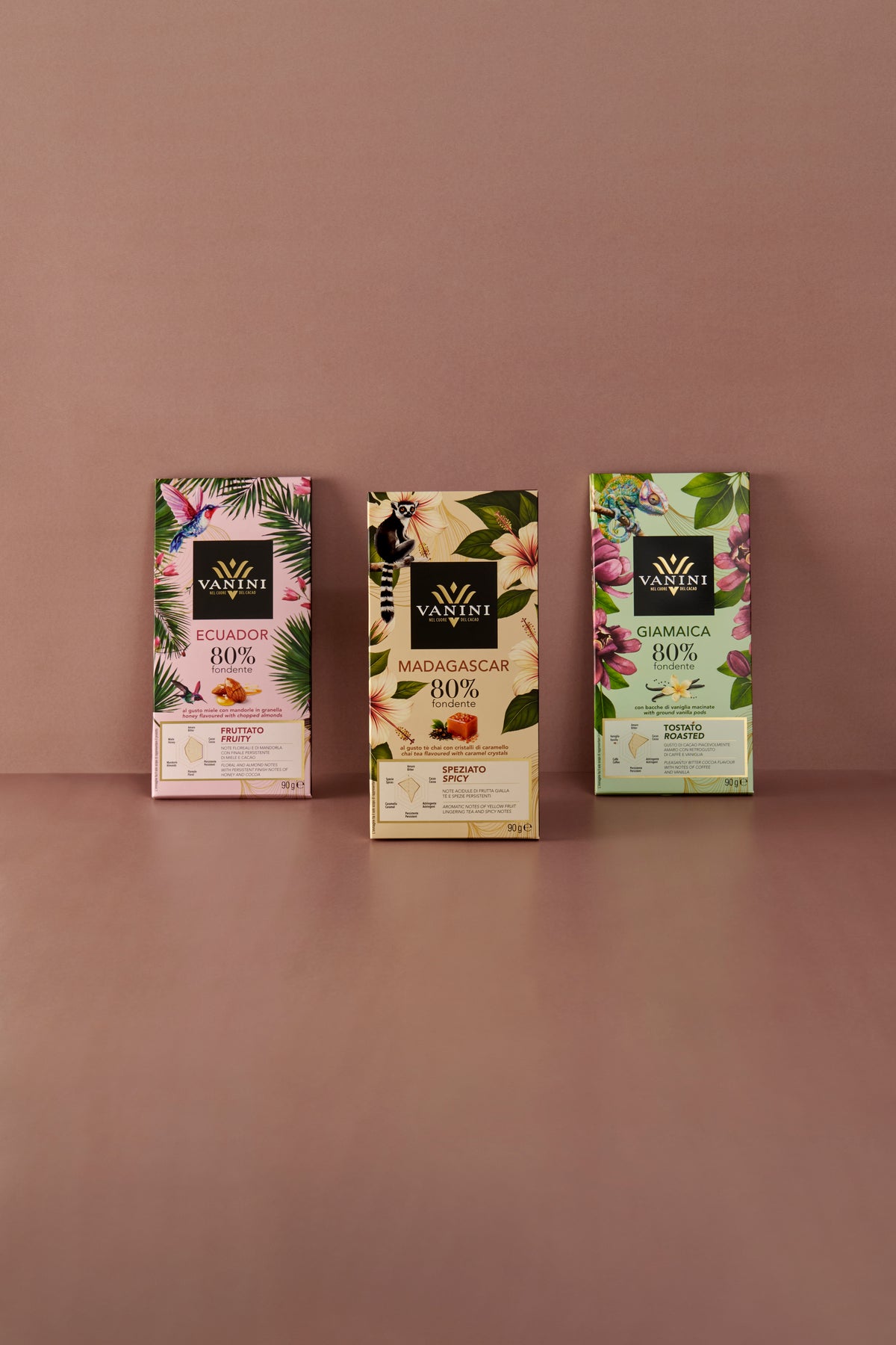 Tre tavolette di cioccolato fondente Vanini Monorigine 80% disposte su sfondo rosa. Da sinistra: Ecuador dal profilo aromatico fruttato, Madagascar dal profilo speziato e Giamaica dal profilo tostato, con confezioni decorate da motivi tropicali, fiori e animali esotici.