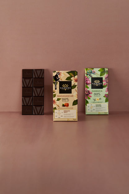 Tre tavolette di cioccolato fondente Vanini Monorigine 80% disposte su sfondo rosa. Da sinistra: Ecuador (scartato) dal profilo aromatico fruttato, Madagascar dal profilo speziato e Giamaica dal profilo tostato, con confezioni decorate da motivi tropicali, fiori e animali esotici.