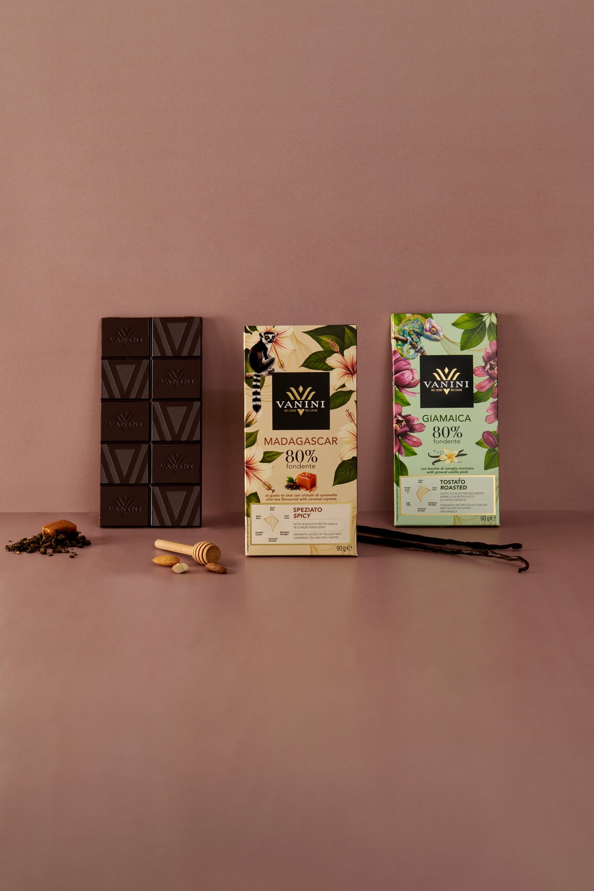 Tavolette Vanini Monorigine 80% fondente Madagascar e Giamaica con una tavoletta di cioccolato fondente in primo piano e ingredienti che richiamano i profili aromatici, su sfondo rosa.