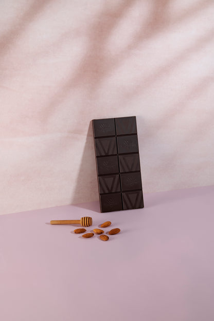 Tavoletta di cioccolato fondente Vanini Ecuador 80% senza confezione, disposta verticalmente su una superficie rosa tenue. In primo piano sono presenti mandorle intere e un spargimiele in legno, ingredienti che richiamano le note di miele e mandorla della ricetta. La finitura scura del cioccolato e l'incisione del logo Vanini risaltano sullo sfondo luminoso e minimale.