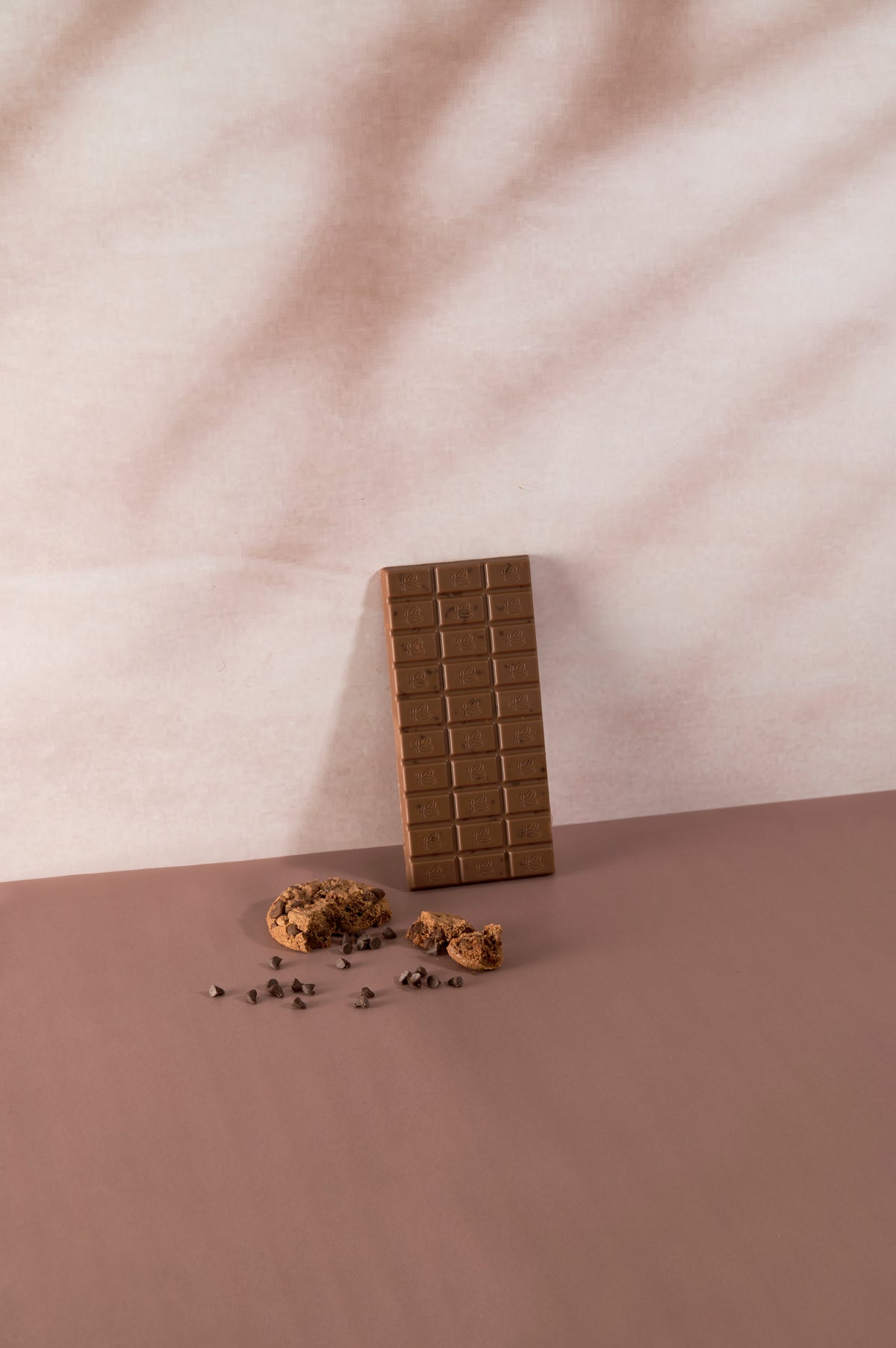 Tavoletta di cioccolato al latte Vanini con cookies e gocce di cioccolato, fotografata accanto a biscotti croccanti e pepite di cioccolato su set minimal dai colori pastello.