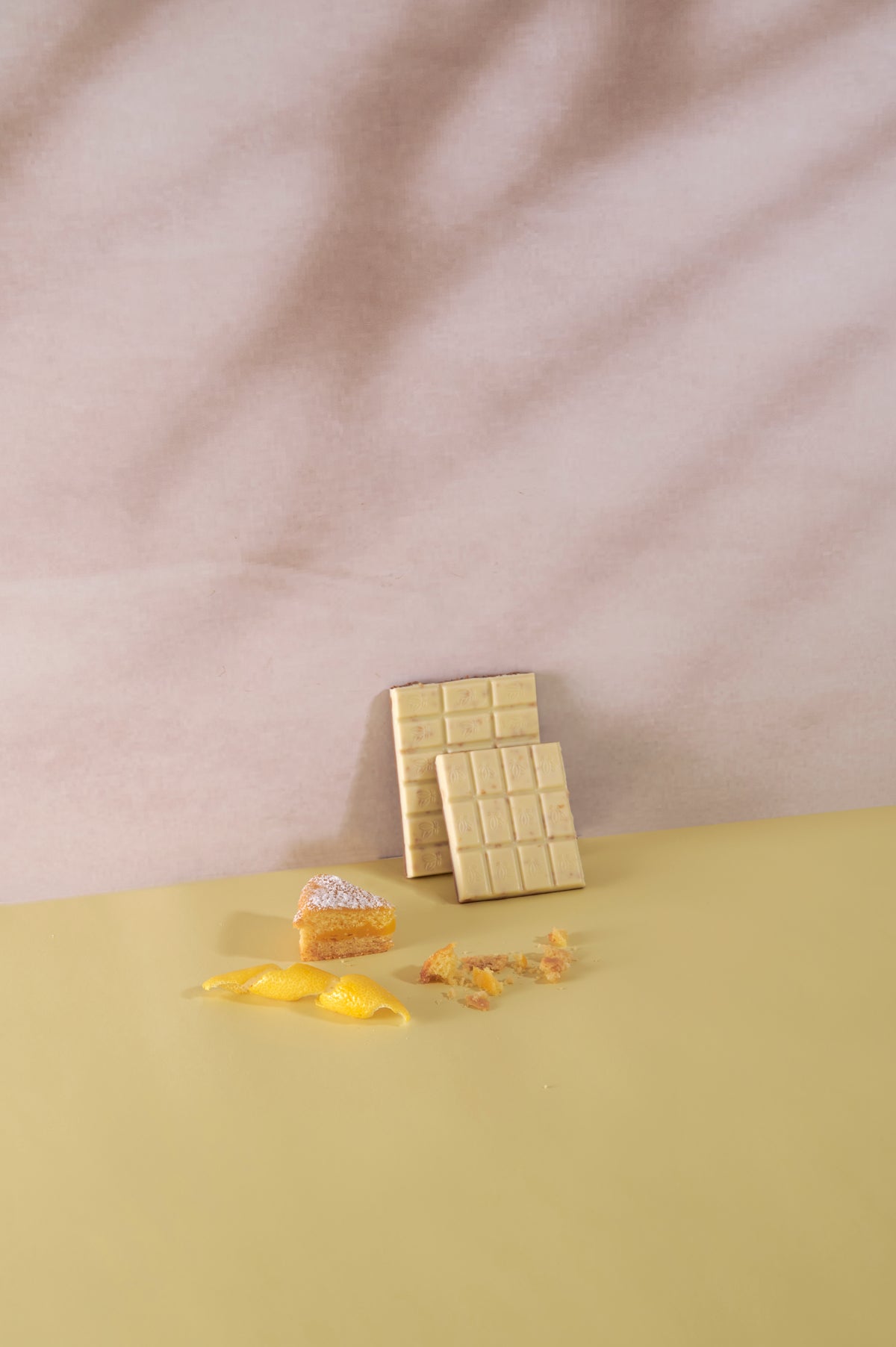 Tavoletta di cioccolato bianco Vanini Tasting Experience Double Torta al Limone accompagnata da una fetta di lemon cake, scorza di limone e briciole di dolce su sfondo giallo tenue.
