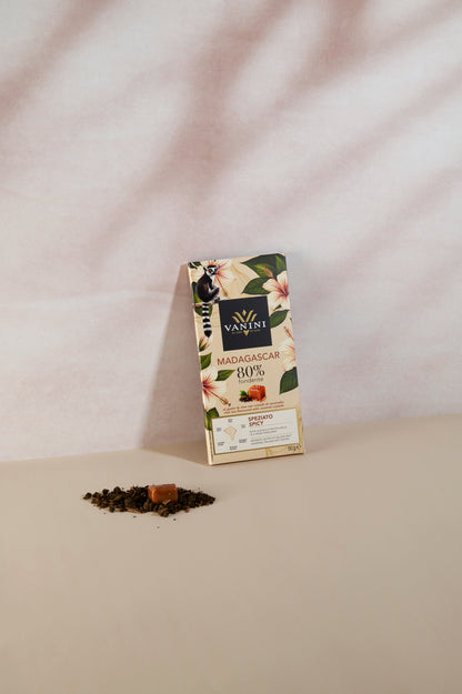 Tavoletta di cioccolato fondente Vanini Madagascar 80% con tè chai e cristalli di caramello, confezione illustrata con fiori tropicali e lemure del Madagascar. Il prodotto è fotografato in verticale su una superficie beige con sfondo chiaro dalle ombre morbide, in uno stile elegante e minimale.