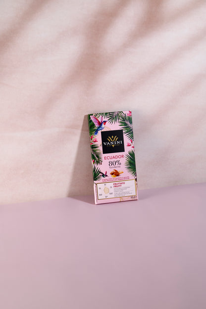 Confezione della tavoletta Vanini Ecuador 80% fondente con miele e mandorle in granella, caratterizzata da una grafica tropicale con foglie verdi, fiori rosa e colibrì. La confezione è posizionata in verticale su una superficie rosa pastello, con illuminazione morbida che mette in evidenza il design premium e il profilo aromatico fruttato del cioccolato Monorigine Ecuador.